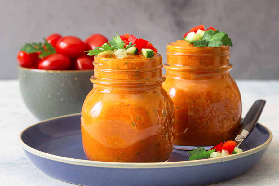 Gazpacho recipe Mood picture: Two screw-top jars filled with red gazpacho arranged on a blue plate, in the background a bowl of tomatoes